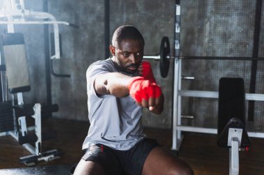 Beden eğitiminde spor. Afrikalı dövüşçü dövüşmeye hazırlanıyor kırmızı boksla yumruk atarken spor sargılarını spor salonunda sarar. Güçlü adam boks antrenmanına hazır yumrukları eğitiyor.