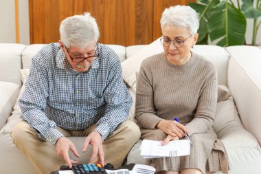 Orta yaşlı yaşlı çift dizüstü bilgisayarları ve kağıt belgeleriyle oturuyor. Yaşlı, olgun bir kadın kağıt faturası okuyor. Ev yöneticiliği yapıyor. Vergileri finanse ediyor.