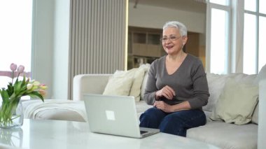 Orta yaşlı mutlu bir kadın dizüstü bilgisayarla oturmuş arkadaşlarıyla görüntülü konuşma yapıyor. Yaşlı ve olgun büyükanneme gülüp yetişkin çocuklarla internette konuşarak eğleniyorum.