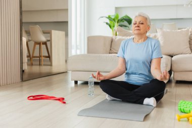 Yoga farkındalığı meditasyonu. Yetişkin bir kadın evde yoga yapıyor. Orta yaşlı kadın nilüfer çiçeğinde oturmuş yoga minderinde meditasyon yaparken poz veriyor. Yaşlı orta yaşlı kadın nefes egzersizi yapıyor.