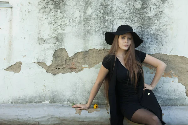 Beautiful young woman in a black dress and a black hat against the old wall — Stock Photo, Image