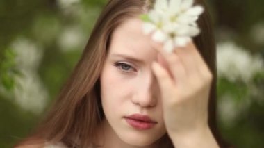 Uzun dalgalı saçlarında çiçekler olan bir kadın çiçek açan elma ağacının yanında rahatlar.