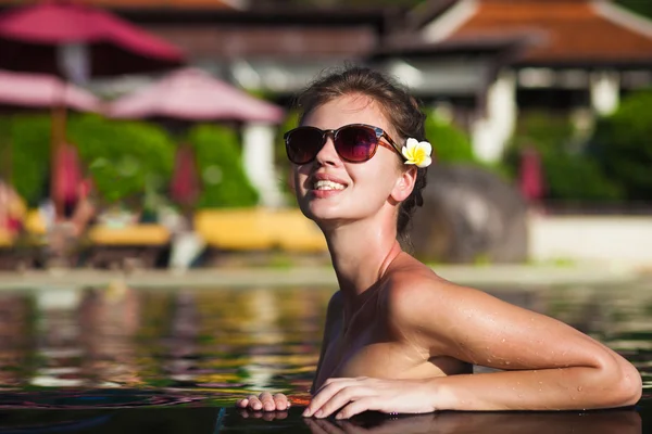 Jolie jeune femme en lunettes de soleil avec fleur dans les cheveux dans la piscine de luxe — Photo