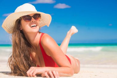 Uzun saçlı, bikinili ve hasır şapkalı bir kadın tropik plajda yatıyor.