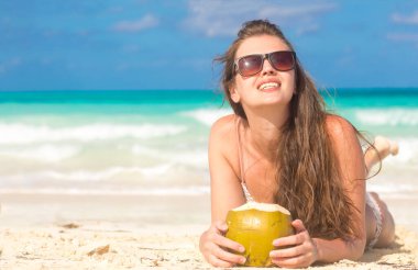 Hasır şapkalı, güneş gözlüklü, kumsalda hindistan cevizli, gülümseyen genç bir kadın.