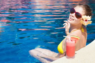 Beautiful young woman in sunglasses with flower in hair smiling in luxury pool clipart