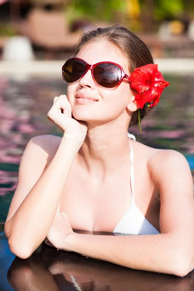 Hermosa mujer joven en gafas de sol con flor en el pelo sonriendo en la piscina de lujo —  Fotos de Stock