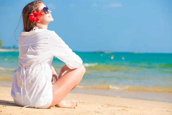 Tillbaka Visa ung långhåriga kvinna i vit skjorta och solglasögon med blomma i håret sitter på stranden — Stockfoto