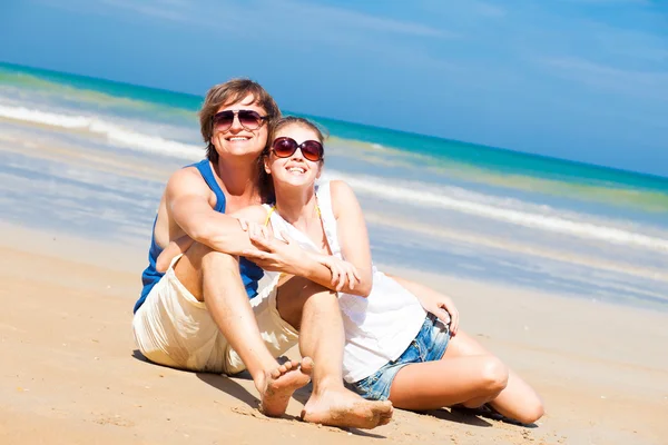 Joyeux jeune couple en vêtements lumineux dans des lunettes de soleil assis sur la plage. les pouces levés — Photo