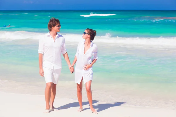 Ritratto di una giovane coppia felice in occhiali da sole in abiti bianchi che cammina e si tiene per mano sulla spiaggia tropicale — Foto Stock