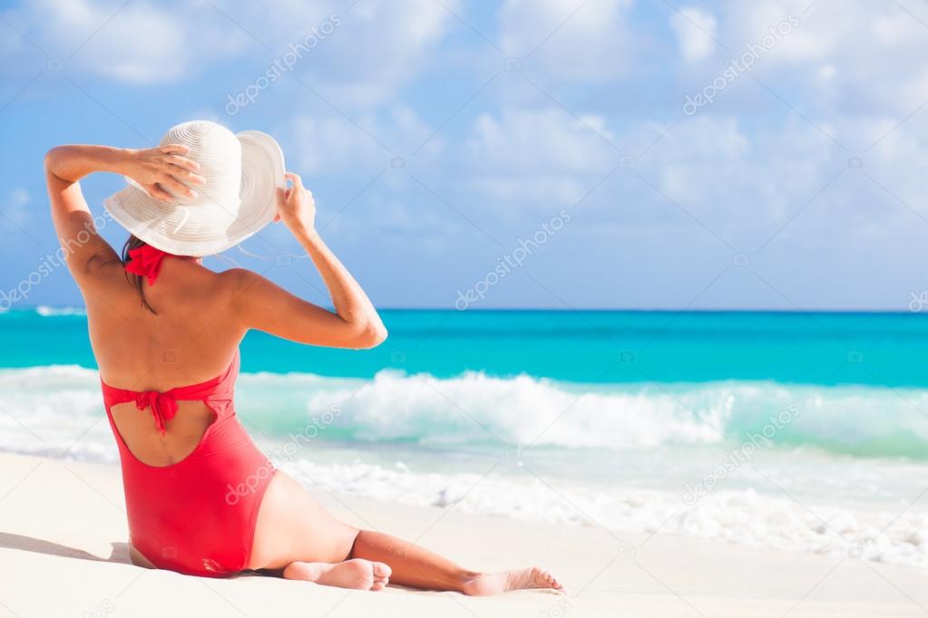 back view of woman in red swim suit and straw hat sitting on tropical beach
