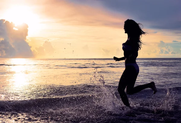大大咧咧的女人，在夕阳下运行在沙滩上。度假的活力健康生活概念。水滴 — 图库照片