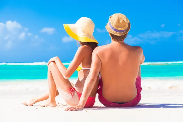 Rückansicht eines glücklichen jungen kaukasischen Paares in Hüten, das am Strand sitzt — Stockfoto