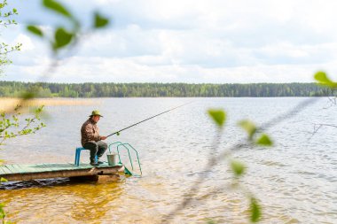 Velikoe Gölü Park Oteli. - Rusya. 22 Mayıs 2021. Elinde olta olan şapkalı bir balıkçı nehrin kıyısında oturur.. 
