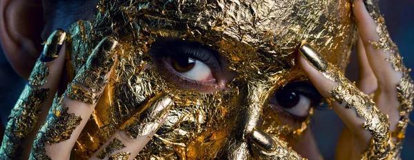 Girl with a mask on her face made of gold leaf. Gloomy studio portrait of a brunette on an abstract background.