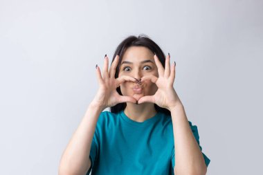 Gri arka planda parmakları izole bir şekilde kalp hareketi gösteren gülümseyen genç bir kadının portresi.