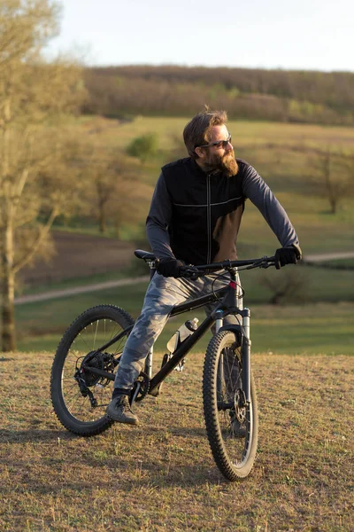 Stock image Sports brutal bearded guy on a modern mountain bike. Cyclist on the green hills in the spring.