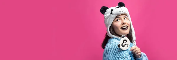 stock image Funny young girl standing with yummy panda-lollipop in her hand and a hat on his head on a pink background.