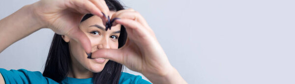 Portrait of a smiling young woman showing heart gesture with her fingers isolated over grey background
