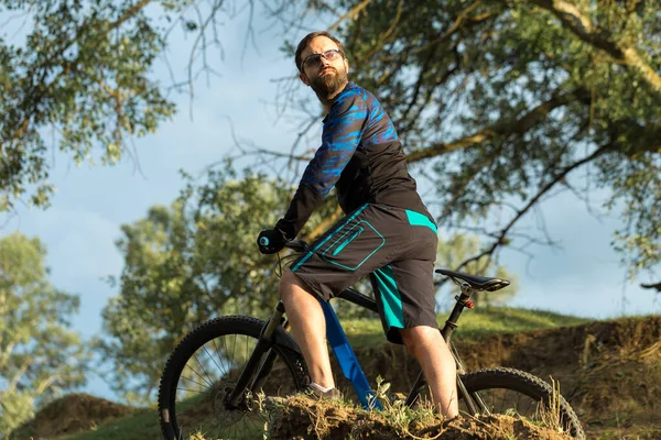 Cyclist in shorts and jersey on a modern carbon hardtail bike with an air suspension fork standing on a cliff against the background of fresh green spring forest