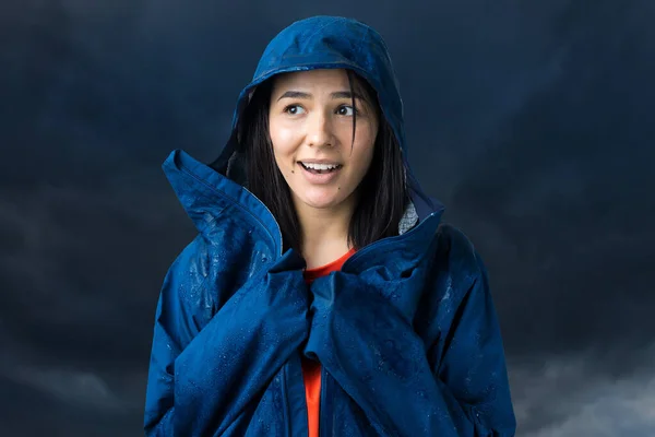 Portrait of a smiling girl dressed in blue raincoat in drops posing with hood on grey background in a studio.