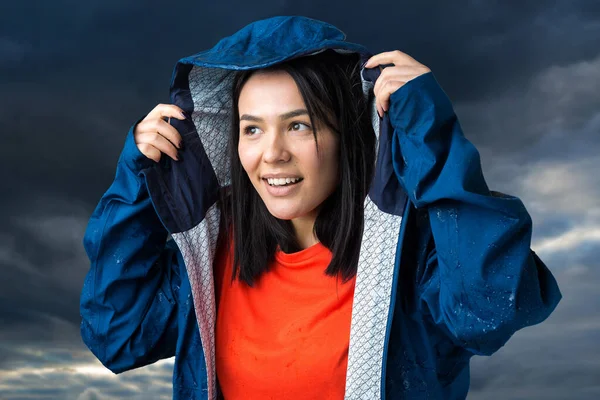 Portrait of a smiling girl dressed in blue raincoat in drops posing with hood on grey background in a studio.