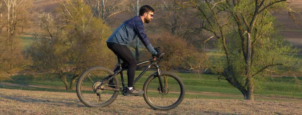 Cyclist in shorts and jersey on a modern carbon hardtail bike with an air suspension fork standing on a cliff against the background of fresh green spring forest