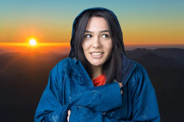 Portrait of a smiling girl dressed in blue raincoat in drops posing with hood against the background of sunset in the mountains.