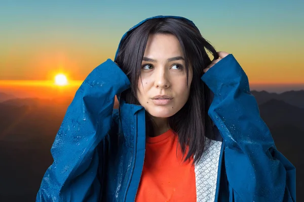 Portrait of a smiling girl dressed in blue raincoat in drops posing with hood against the background of sunset in the mountains.