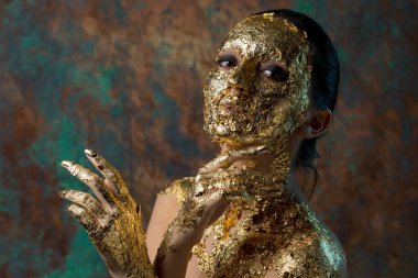 Yüzünde altın yapraktan yapılmış bir maske olan bir kız. Soyut bir arka planda esmer bir kadının kasvetli stüdyo portresi.