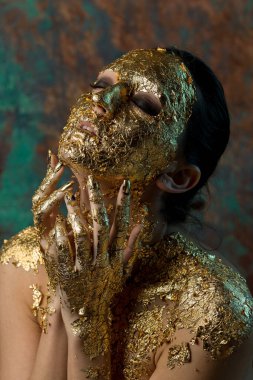Yüzünde altın yapraktan yapılmış bir maske olan bir kız. Soyut bir arka planda esmer bir kadının kasvetli stüdyo portresi.