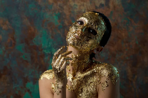 Girl with a mask on her face made of gold leaf. Gloomy studio portrait of a brunette on an abstract background.