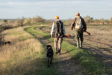 Kamuflaj kıyafetli bir avcı elinde silahla sazlıklar boyunca yürüyor..