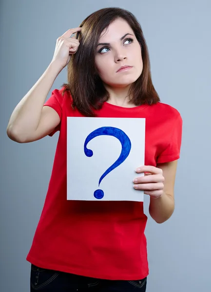 Thoughtful girl in a red T-shirt on a gray background holding a question mark — Stock Photo, Image