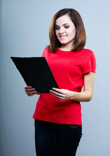 Beautiful girl in a red T-shirt and jeans holding a folder and looking at it on a gray background — Stock Photo, Image