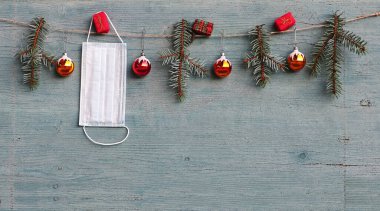 Corona ve Christmas. Noel ağacı topları, köknar dalları ve hediye paketlerinin yanında koruyucu bir maske asılı.