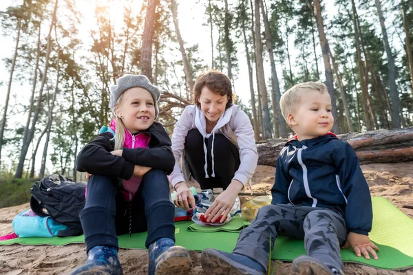 Fiatal, fehér, gyönyörű anya élvezi a pikniken való étkezést két aranyos kisgyerekkel a fenyőerdőben a meleg őszi napon. Családi természet szabadtéri kikapcsolódás életmód — Stock Fotó