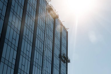 Camdan metal ön cephe şantiyesi olan yüksek kulenin duvarının yanındaki askıya alınmış inşaat beşiği. Şehir geliştirme mühendisliği geçmişi. Güneş gökyüzünde parlıyor