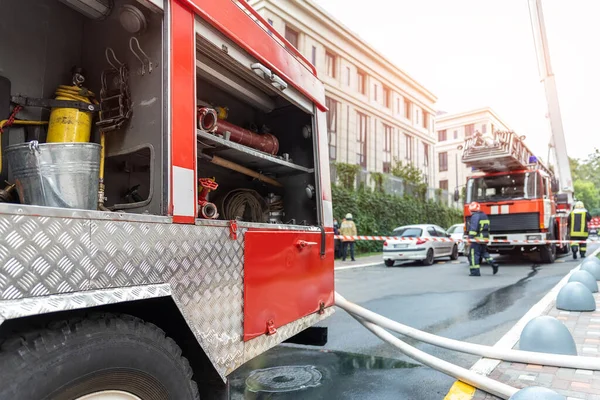 Πυροσβέστες φορώντας στολή στέκεται πίσω από ταινία φωτιά και πολλά πυροσβεστικά οχήματα με σκάλα κατά λάθος σε πύργο highrise κατοικιών ή κτίριο γραφείων στο κέντρο της πόλης. Επείγουσα διάσωση σε περίπτωση καταστροφής — Φωτογραφία Αρχείου