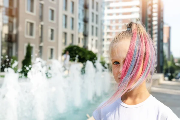 Linda adorable niña caucásica con tiza pastel multicolor pintado pelo rubio teñido en la calle de la ciudad con fondo de fuente. Mujer niño retrato rosa colorido peinado sonriendo al aire libre — Foto de Stock