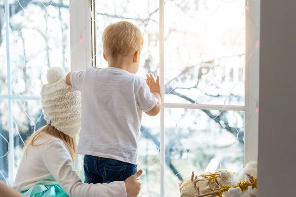 Due simpatici adorabili piccoli bambini biondi cauasiatici fratelli rimangono vicino alla finestra e guardando fuori in attesa di neve, meraviglie e miracolo durante le vacanze di Natale. Resta a casa al covid-19, isolamento pandemico. — Foto Stock