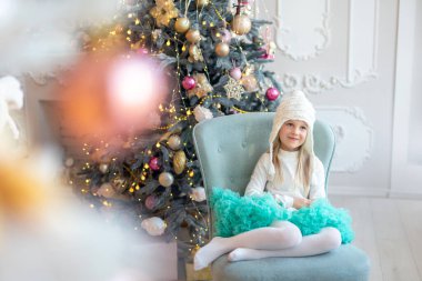 Şirin, sevimli, beyaz, sarışın, küçük bir kız çocuğu Noel ağacının yanındaki sandalyesinde oturuyor ve Noel mucizelerini ve mucizelerini hayal ediyor. Olumlu düşünceli çocuk portresi Yeni yıl arifesi tatili