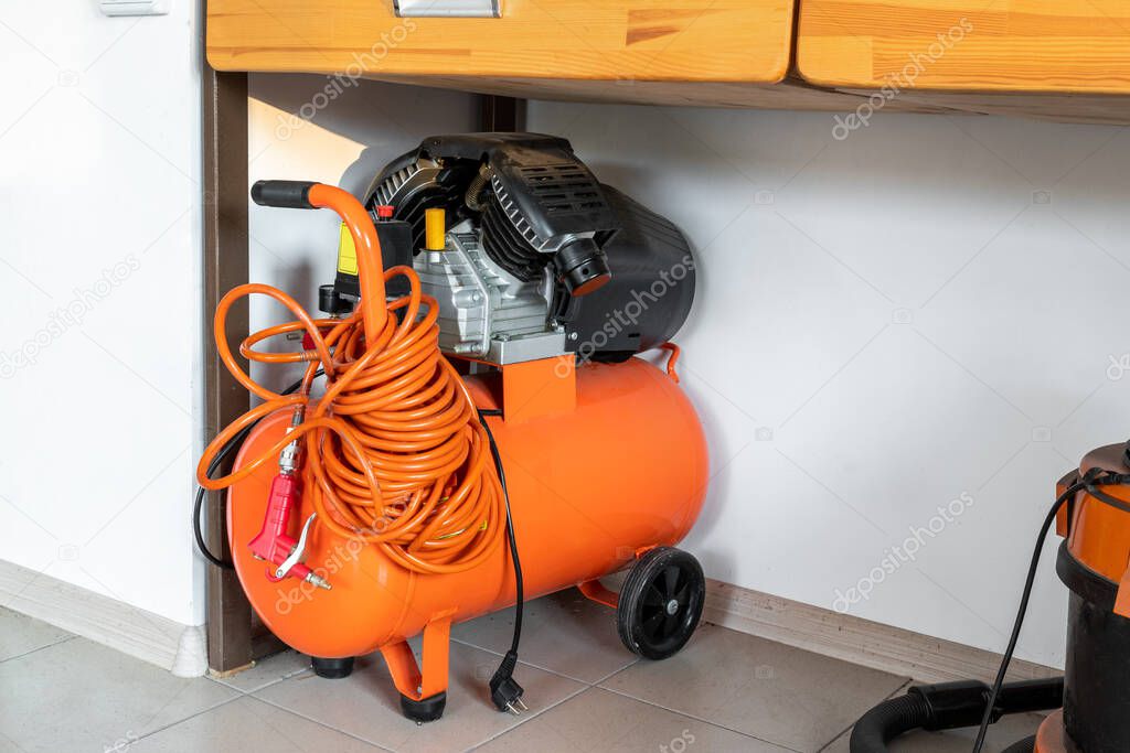 Orange small portable industrial power air compressor with coil hose and pneumatic gun at home warehouse garage under wooden workbench. Electric tools and equipment.