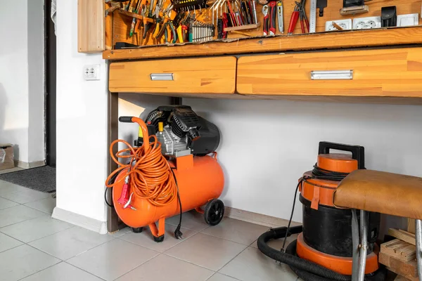 Orange small portable industrial power air compressor with coil hose and pneumatic gun and vacuum cleaner at home warehouse garage under wooden workbench. Electric tools and equipment.