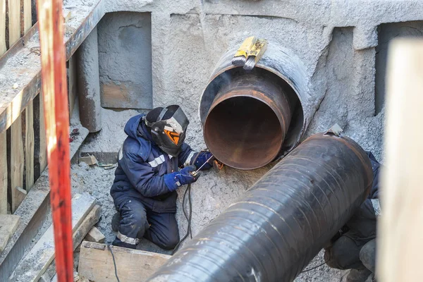 Professional welder welding water or gas steel pipeline in protective trench and mask at construction site pit. City underground utilitites renewal replacement. Emergency pipe break leak elimination