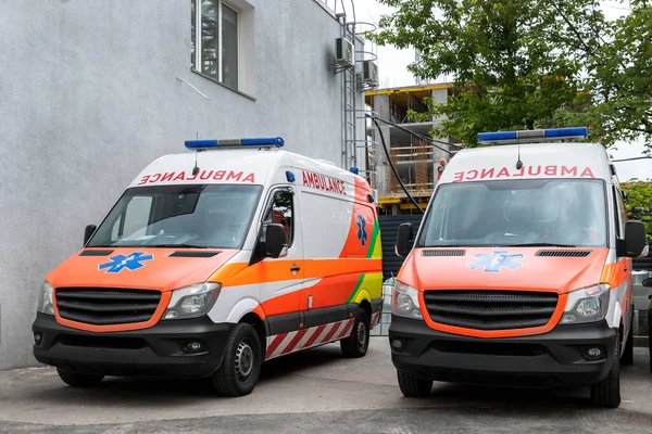 Két modern fehér és narancssárga mentőautó áll a kórház épülete előtt. Mentősök a város utcájában. Egészségügyi ellátás és sürgős orvosi ellátás koncepciója — Stock Fotó