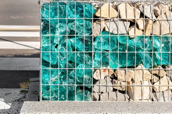 Detalhe de close-up da nova cerca de gabião bonito moderno com gaiola de metal preenchido por pedra esmagada e pedras de vidro azul artificial quebrado. Cidade rua proteção pavimento rodoviário — Fotografia de Stock