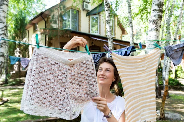 Candid real life portrait of young adult beautiful attractive caucasian woman hanging up fresh washed family clothes on birch tree clothesline with pins at home yard on bright sunny day outdoors — Stock Photo, Image