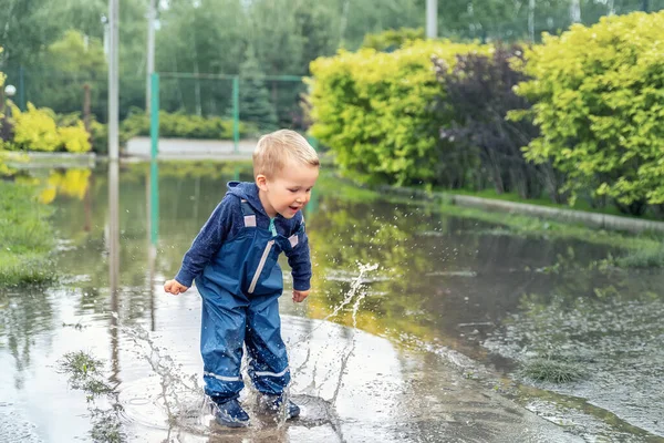 Pequeño niño pequeño rubio caucásico juguetón disfrutar de divertirse jugando saltar en charco sucio con pantalones azules impermeables y botas de lluvia de goma en casa patio calle al aire libre. Concepto de infancia feliz — Foto de Stock