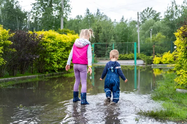 Vista trasera de par de dos lindos niños caucásicos rubios hermano y hermana disfrutan de divertirse jugando saltar en charco sucio usando pantalones impermeables azules y botas de lluvia de goma en la calle al aire libre — Foto de Stock